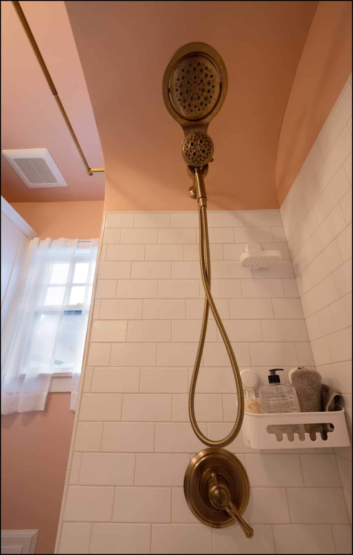 A gold-colored showerhead with a handheld attachment is mounted on a tiled wall in a bathroom with peach-colored walls and a window with sheer curtains.