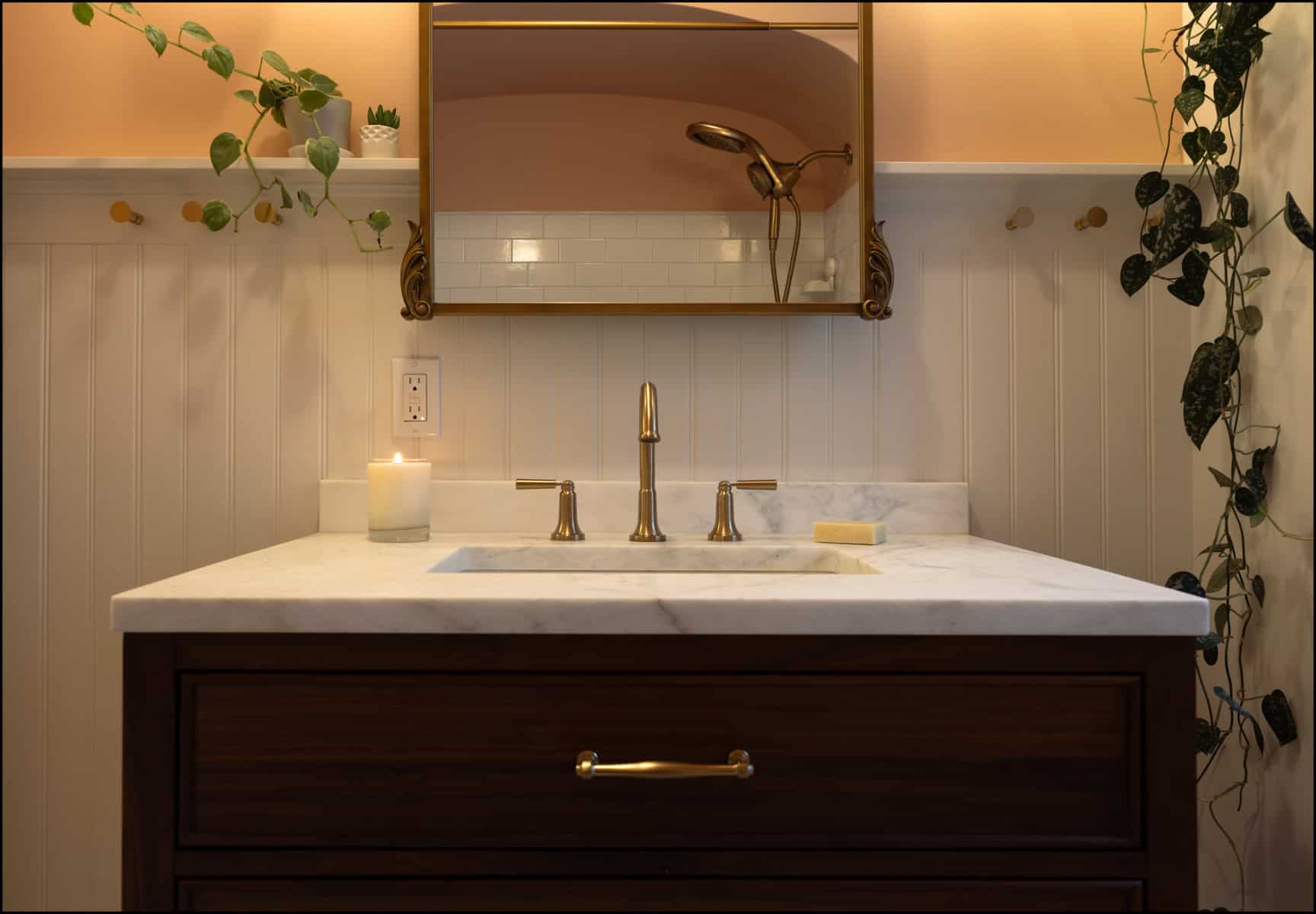 A bathroom sink with a marble countertop, brass faucet, lit candle, bar of soap, mirror, and hanging plants against a pale paneled wall.