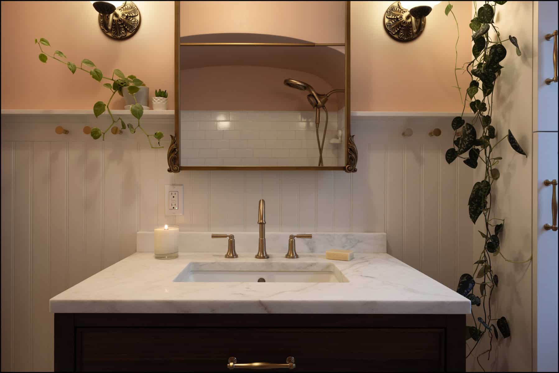 A bathroom vanity with a marble countertop, gold fixtures, a soap bar, a lit candle, wall plants, and a mirror reflecting a showerhead against white tile.