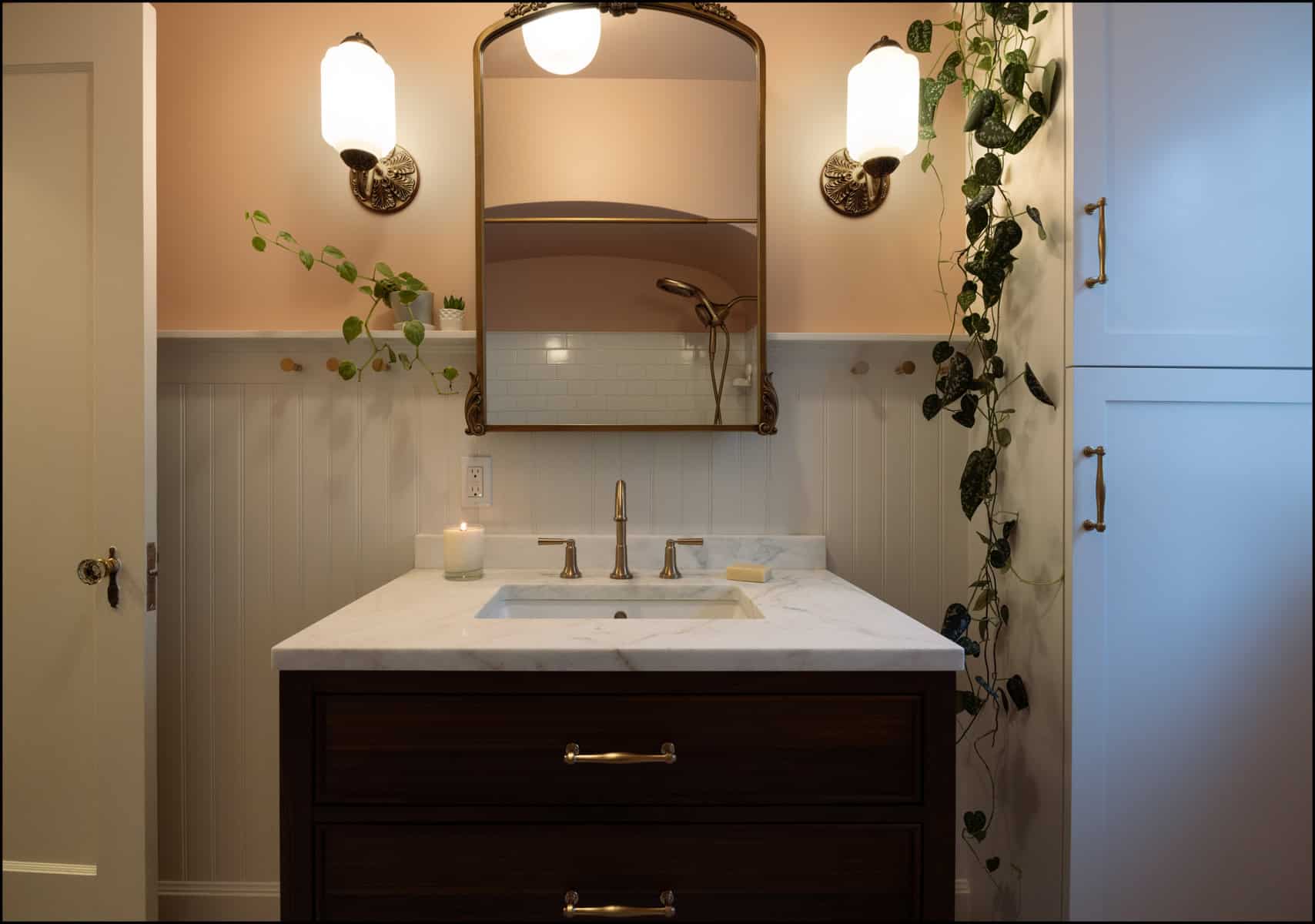 A bathroom vanity with a marble countertop, gold faucet, rectangular mirror, wall sconces, trailing plants, and white cabinetry.