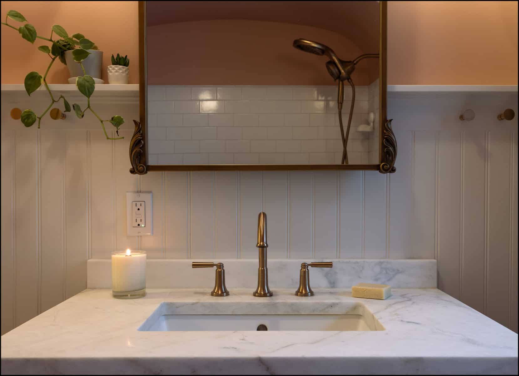 A bathroom sink with a marble countertop, brass faucet, lit candle, bar of soap, potted plants, and a mirror reflecting a showerhead and tiled wall.