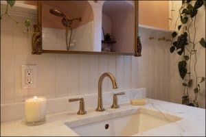 A bathroom sink with gold fixtures, a lit candle, a bar of soap, and a wall mirror. Plants hang on the wall and a shower is visible in the background.