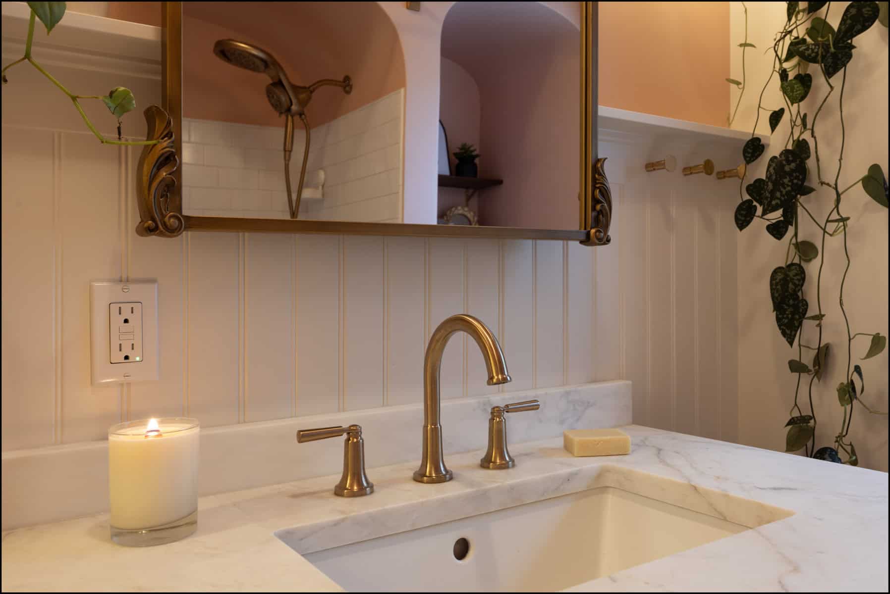 A bathroom sink with gold fixtures, a lit candle, a bar of soap, and a wall mirror. Plants hang on the wall and a shower is visible in the background.
