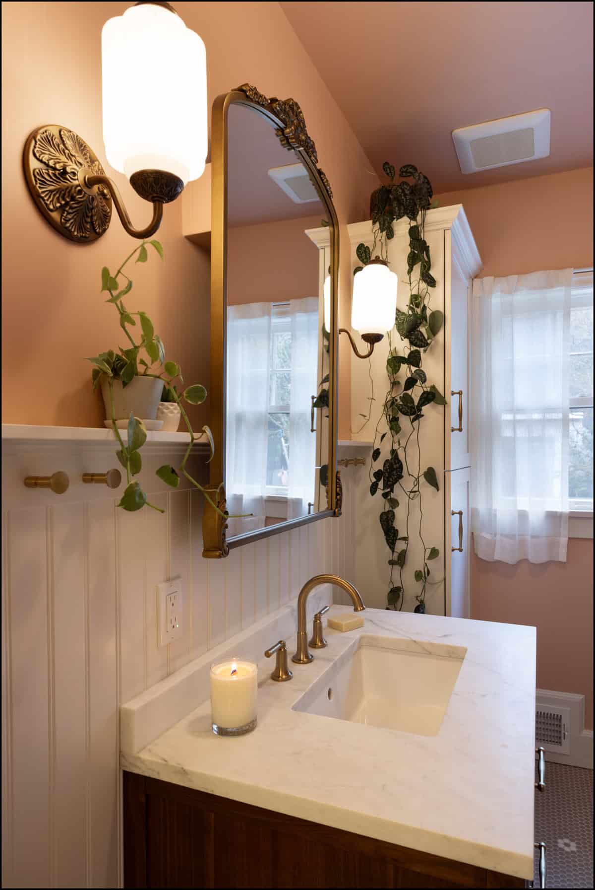A bathroom with a marble sink, gold faucet, wall mirror, lit candle, wall-mounted lights, potted plant, and trailing vines on peach and white walls near a window with sheer curtains.