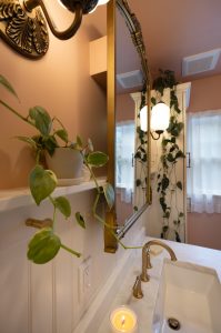 A bathroom with a gold-framed mirror, wall-mounted lamp, potted plant, lit candle on the sink, and trailing vine plants near a window with sheer curtains.
