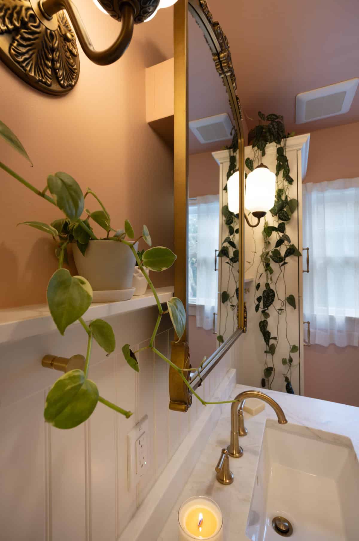 A bathroom with a gold-framed mirror, wall-mounted lamp, potted plant, lit candle on the sink, and trailing vine plants near a window with sheer curtains.