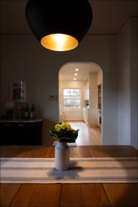 A small vase with yellow flowers sits on a striped runner atop a wooden dining table, with a softly lit kitchen visible through an arched doorway in the background.