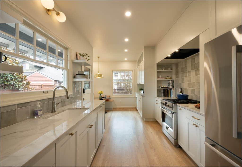 Modern kitchen with white cabinets, marble countertops, stainless steel appliances, wooden floor, and large window overlooking a side yard.