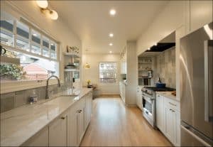 Modern kitchen with white cabinets, marble countertops, stainless steel appliances, wooden floor, and large window overlooking a side yard.