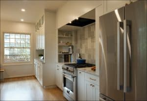 Modern kitchen with stainless steel refrigerator, gas stove, oven, white cabinets, tiled backsplash, wooden floor, and a window letting in natural light.