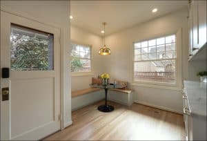 Small, bright breakfast nook with a built-in wooden bench, round black table, yellow flowers in a vase, and large windows overlooking a fenced yard.