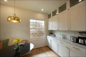 Bright kitchen with white cabinets, marble countertops, a black round table with a vase of yellow flowers, and a window overlooking trees. Coffee maker and jars are on the counter.