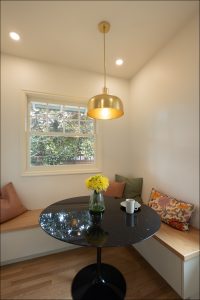 A small dining nook with a round black table, a vase of yellow flowers, a mug on a saucer, built-in wooden benches with cushions, and a brass pendant light above.