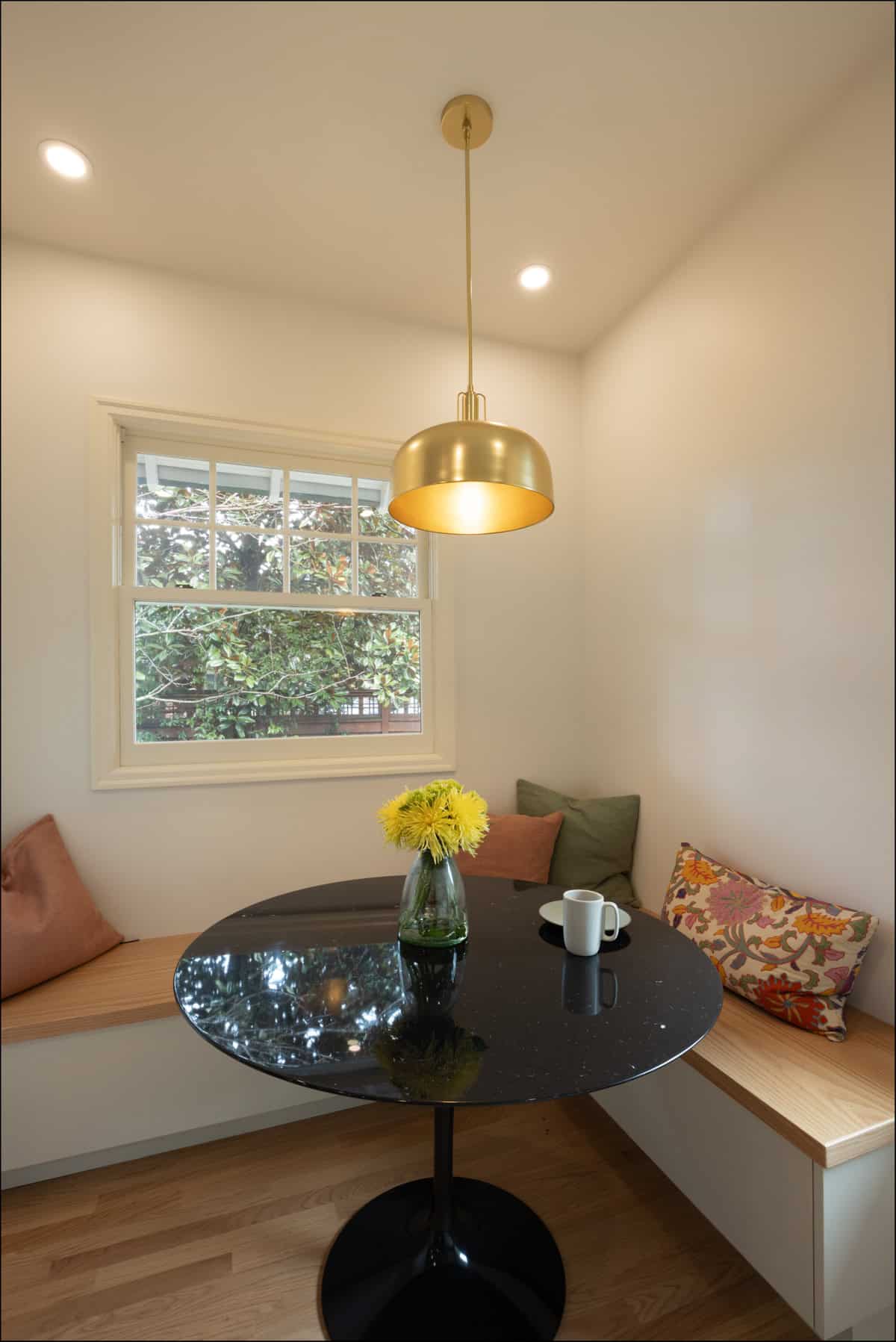 A small dining nook with a round black table, a vase of yellow flowers, a mug on a saucer, built-in wooden benches with cushions, and a brass pendant light above. A small dining nook with a round black table, a vase of yellow flowers, a mug on a saucer, built-in wooden benches with cushions, and a brass pendant light above.