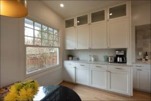 A bright kitchen with white cabinets, a coffee maker, glass jars on the counter, and a large window overlooking a fenced yard.