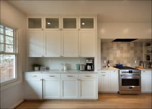 Modern kitchen with white cabinets, marble countertop, coffee maker, and glass storage jars; stove and tiled backsplash visible on the right, large window on the left.