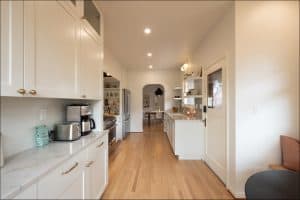 A modern galley kitchen with white cabinets, marble countertops, wooden floor, stainless steel appliances, and shelves; a dining area is visible through an arched doorway.