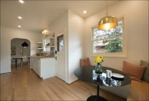 A bright kitchen with light wood floors, a small black dining table with two chairs, a vase of yellow flowers, and a window with cushions on either side.