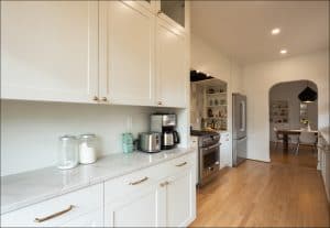 Modern kitchen with white cabinets, marble countertops, coffee maker, stove, and refrigerator; wood floors and view into dining area with shelves and a pendant light.