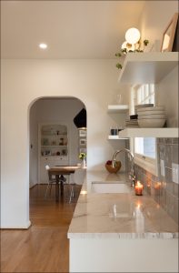 Modern kitchen with marble countertop, open shelves holding dishes, a lit candle by the sink, and a view of a dining area through an arched doorway.