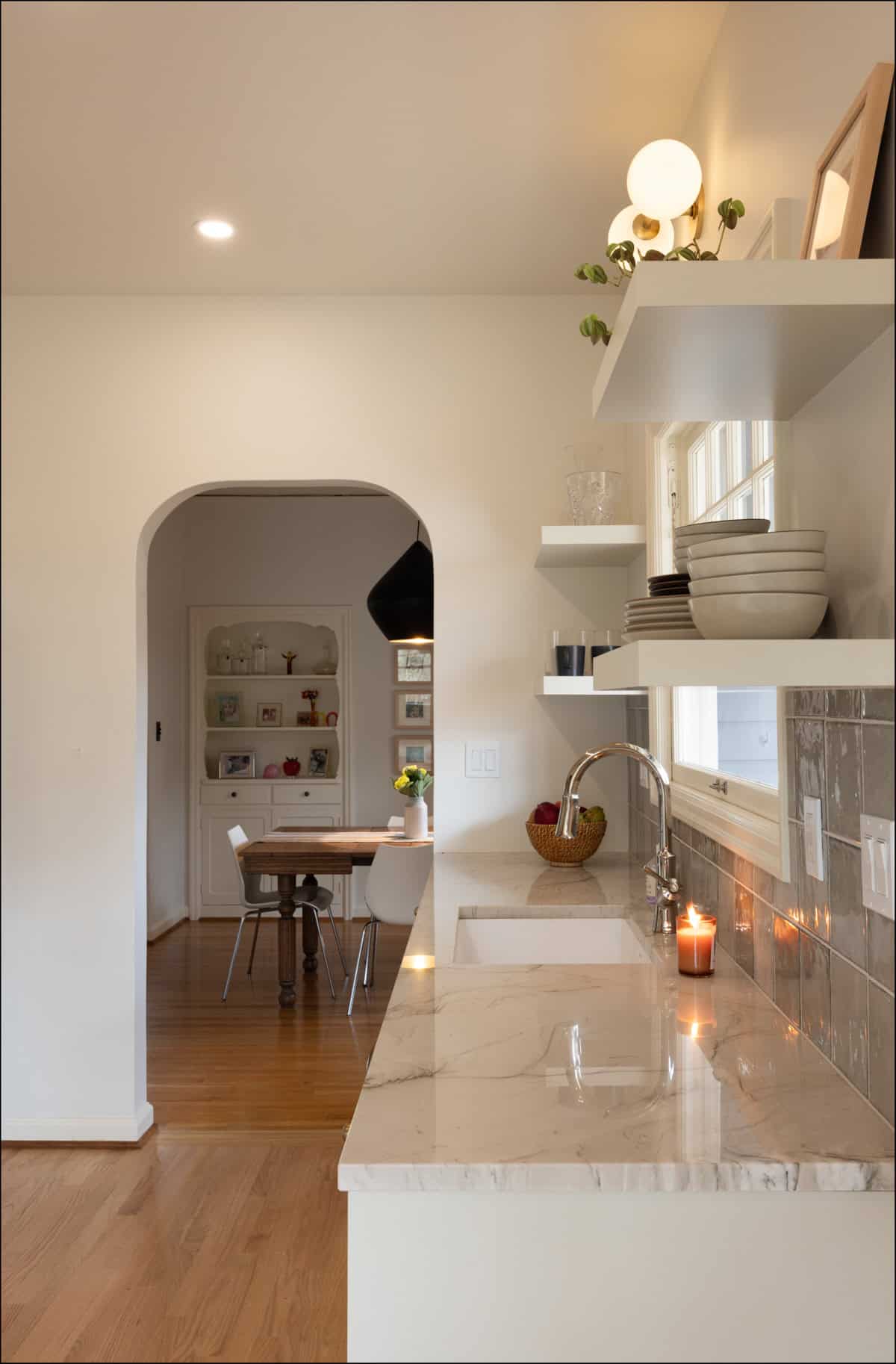 Modern kitchen with marble countertop, open shelves holding dishes, a lit candle by the sink, and a view of a dining area through an arched doorway. Modern kitchen with marble countertop, open shelves holding dishes, a lit candle by the sink, and a view of a dining area through an arched doorway.