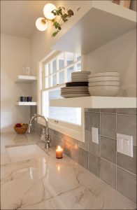 A modern kitchen with a marble countertop, ceramic dishes on open shelves, a lit candle near the sink, and a window above grey-tiled backsplash.