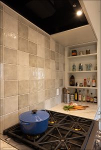 A blue Dutch oven sits on a gas stove in a kitchen with gray tiled backsplash; shelves hold bottles, jars, and kitchen tools.