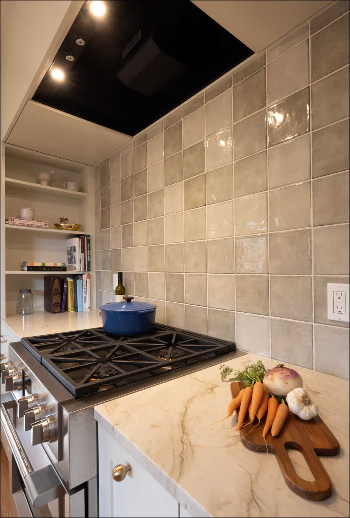 A marble kitchen countertop with carrots, garlic, and a turnip on a cutting board beside a gas stove with a blue pot; tiled backsplash and shelves in the background. A marble kitchen countertop with carrots, garlic, and a turnip on a cutting board beside a gas stove with a blue pot; tiled backsplash and shelves in the background.