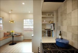 A kitchen with a blue Dutch oven on the stove, open shelving with books and decor, and a dining nook with a round table, bench seating, and flowers in a vase.