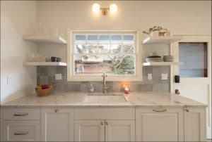 Modern kitchen with white cabinets, marble countertop, open shelves with dishes, a basket of fruit, a lit candle, and a window above the sink letting in natural light.