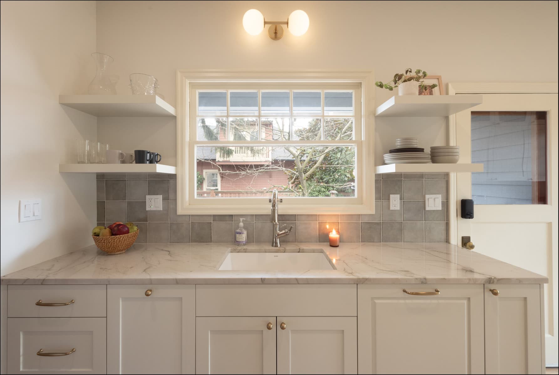 Modern kitchen with white cabinets, marble countertop, open shelves with dishes, a basket of fruit, a lit candle, and a window above the sink letting in natural light. Modern kitchen with white cabinets, marble countertop, open shelves with dishes, a basket of fruit, a lit candle, and a window above the sink letting in natural light.