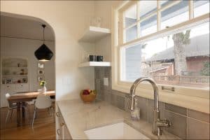 Modern kitchen with marble countertop, stainless steel faucet, open shelves, and a bowl of fruit; dining area with table and chairs visible in the background.