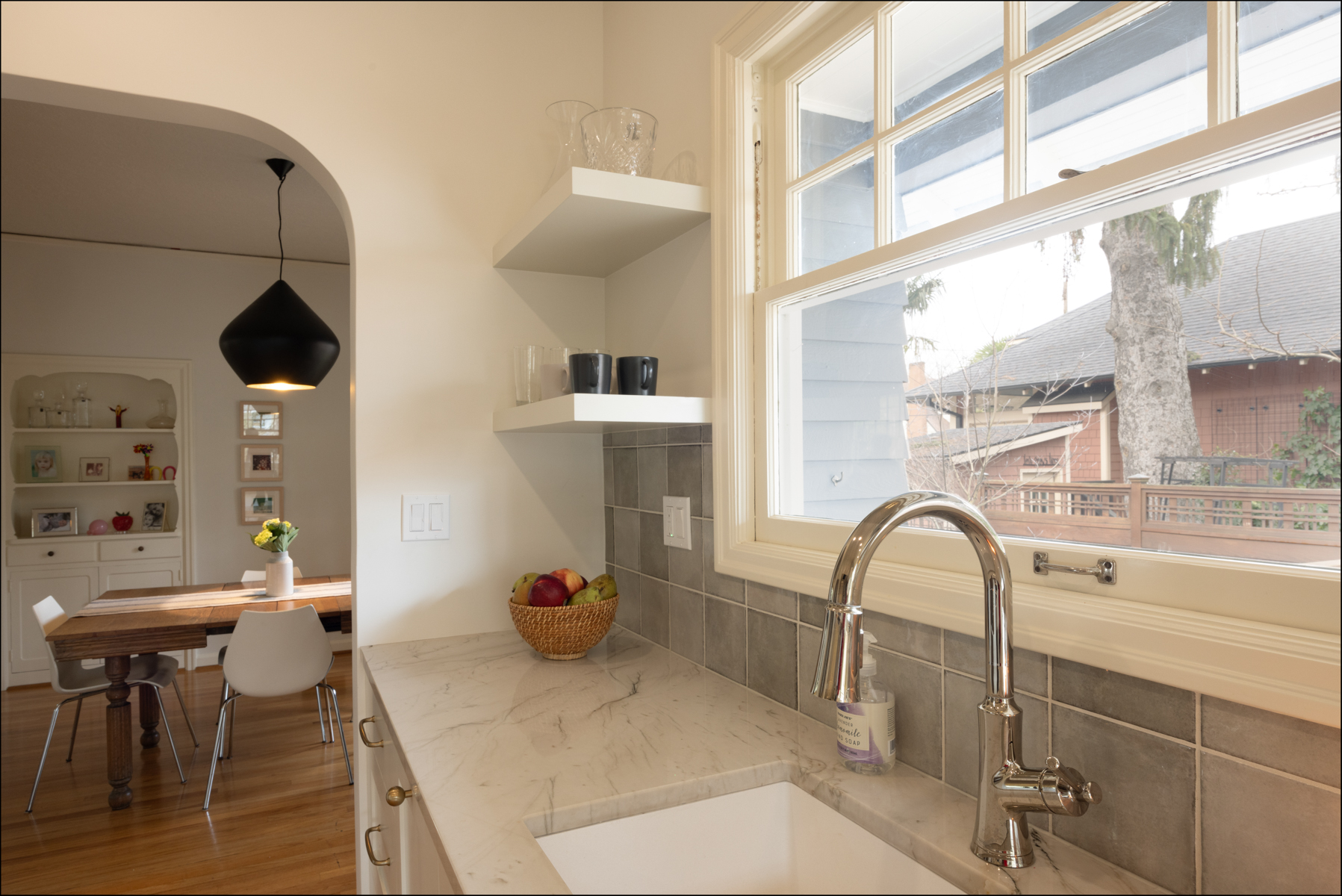 Modern kitchen with marble countertop, stainless steel faucet, open shelves, and a bowl of fruit; dining area with table and chairs visible in the background. Modern kitchen with marble countertop, stainless steel faucet, open shelves, and a bowl of fruit; dining area with table and chairs visible in the background.