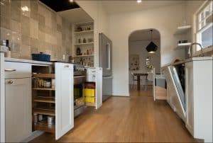 Modern kitchen with light wood floors, several open cabinets, visible pantry shelves, stainless steel fridge, and view into a dining area through an arched doorway.