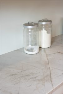 Two glass jars with metal lids, one partially filled and one mostly filled with white powder, sit on a marble countertop.