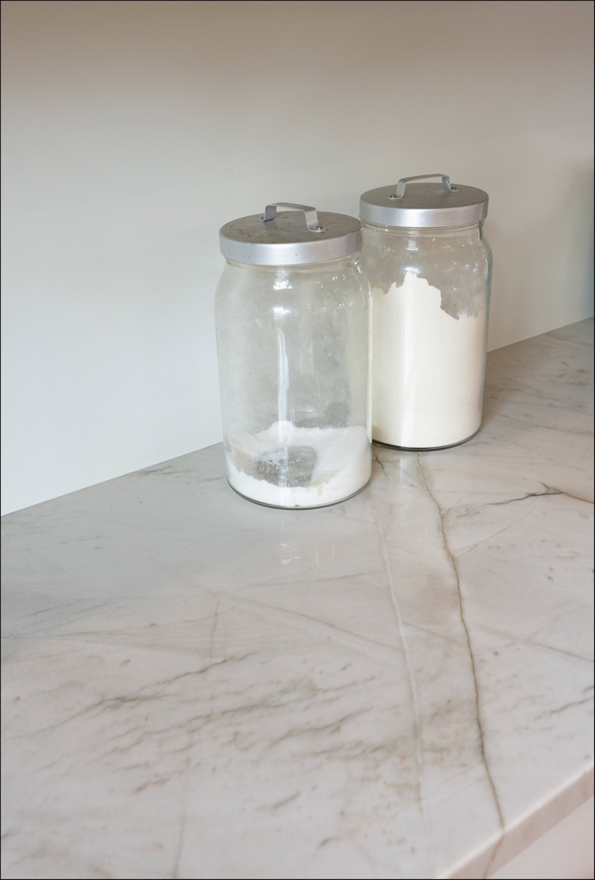 Two glass jars with metal lids, one partially filled and one mostly filled with white powder, sit on a marble countertop. Two glass jars with metal lids, one partially filled and one mostly filled with white powder, sit on a marble countertop.