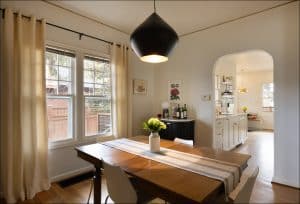 A dining room with a wooden table, four chairs, a striped table runner, and a vase of flowers, adjacent to a kitchen visible through an arched doorway. Natural light fills the space.