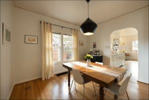 A dining room with a wooden table, four white chairs, a striped table runner, a hanging light fixture, and a view into a bright kitchen through an arched doorway.