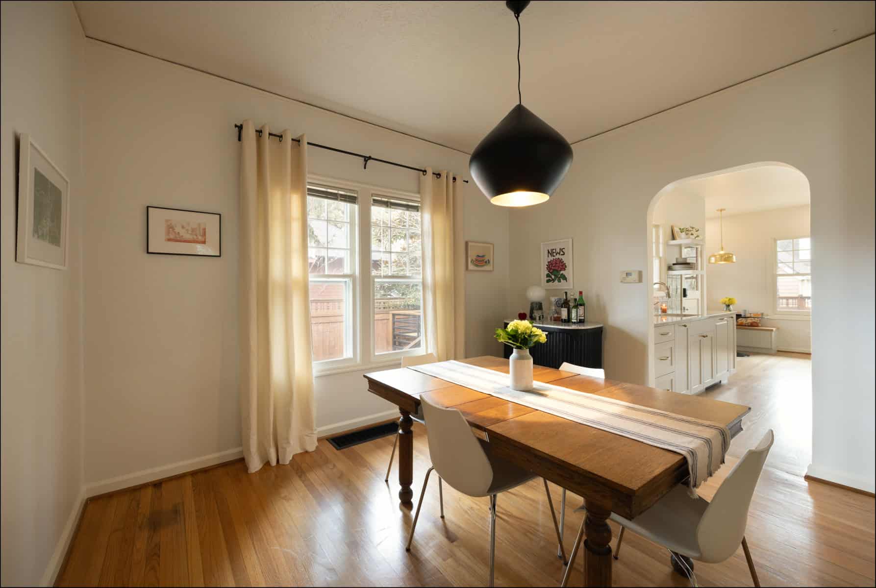A dining room with a wooden table, four white chairs, a striped table runner, a hanging light fixture, and a view into a bright kitchen through an arched doorway. A dining room with a wooden table, four white chairs, a striped table runner, a hanging light fixture, and a view into a bright kitchen through an arched doorway.