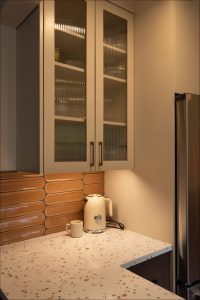 A corner kitchen countertop with a white kettle, a mug, beige terrazzo surface, orange rectangular tile backsplash, glass-fronted upper cabinets, and a stainless steel fridge.