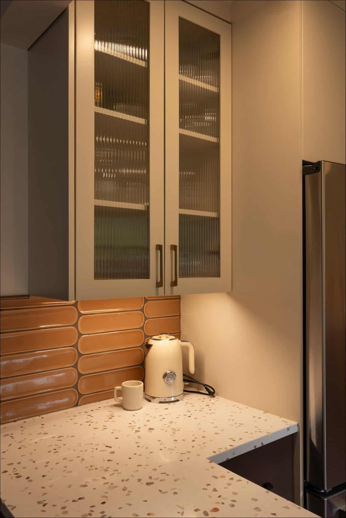 A corner kitchen countertop with a white kettle, a mug, beige terrazzo surface, orange rectangular tile backsplash, glass-fronted upper cabinets, and a stainless steel fridge. A corner kitchen countertop with a white kettle, a mug, beige terrazzo surface, orange rectangular tile backsplash, glass-fronted upper cabinets, and a stainless steel fridge.