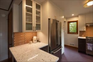 Modern kitchen with white cabinets, stainless steel refrigerator, beige tile backsplash, speckled white countertop, and a window showing greenery outside.