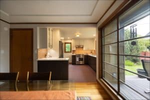 Modern kitchen with white and dark cabinets, marble countertops, and stainless steel appliances, viewed from a dining area next to large windows overlooking a backyard patio.