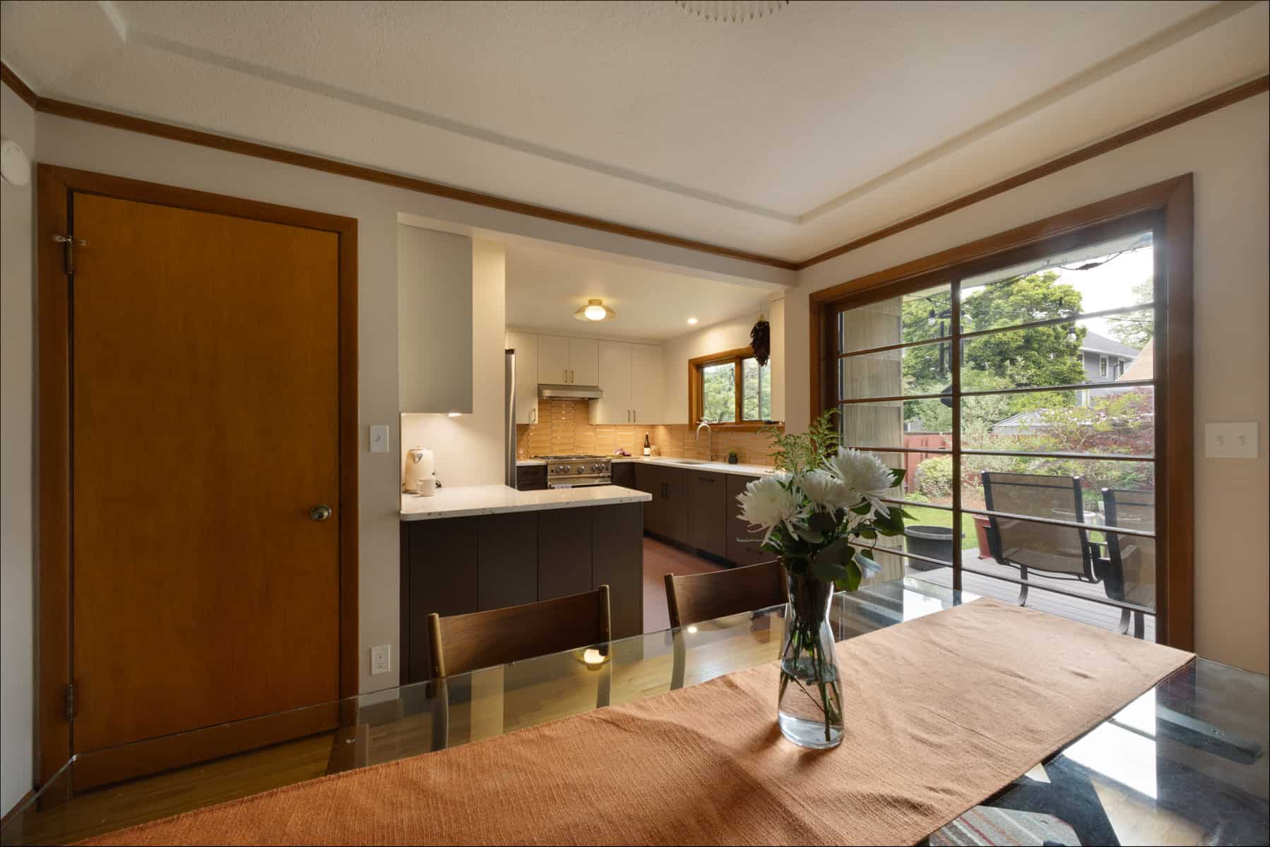 A dining area with a glass table and orange runner, adjacent to a kitchen; large sliding doors open to a deck with greenery outside. A dining area with a glass table and orange runner, adjacent to a kitchen; large sliding doors open to a deck with greenery outside.