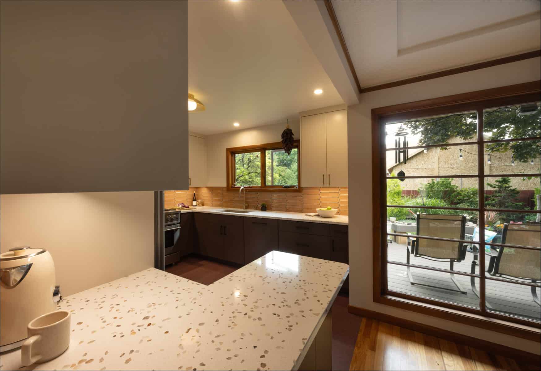 Modern kitchen with white countertops, beige cabinets, and a window overlooking a patio with outdoor seating and trees visible through a large glass door. Modern kitchen with white countertops, beige cabinets, and a window overlooking a patio with outdoor seating and trees visible through a large glass door.
