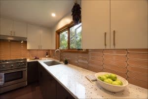 Modern kitchen with stainless steel stove, beige cabinets, brown tile backsplash, white speckled countertop, bowl of green apples, and large window above the sink.