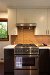 A modern kitchen with a stainless steel gas stove, beige tile backsplash, white upper cabinets, and a countertop with a towel hanging on the oven handle.