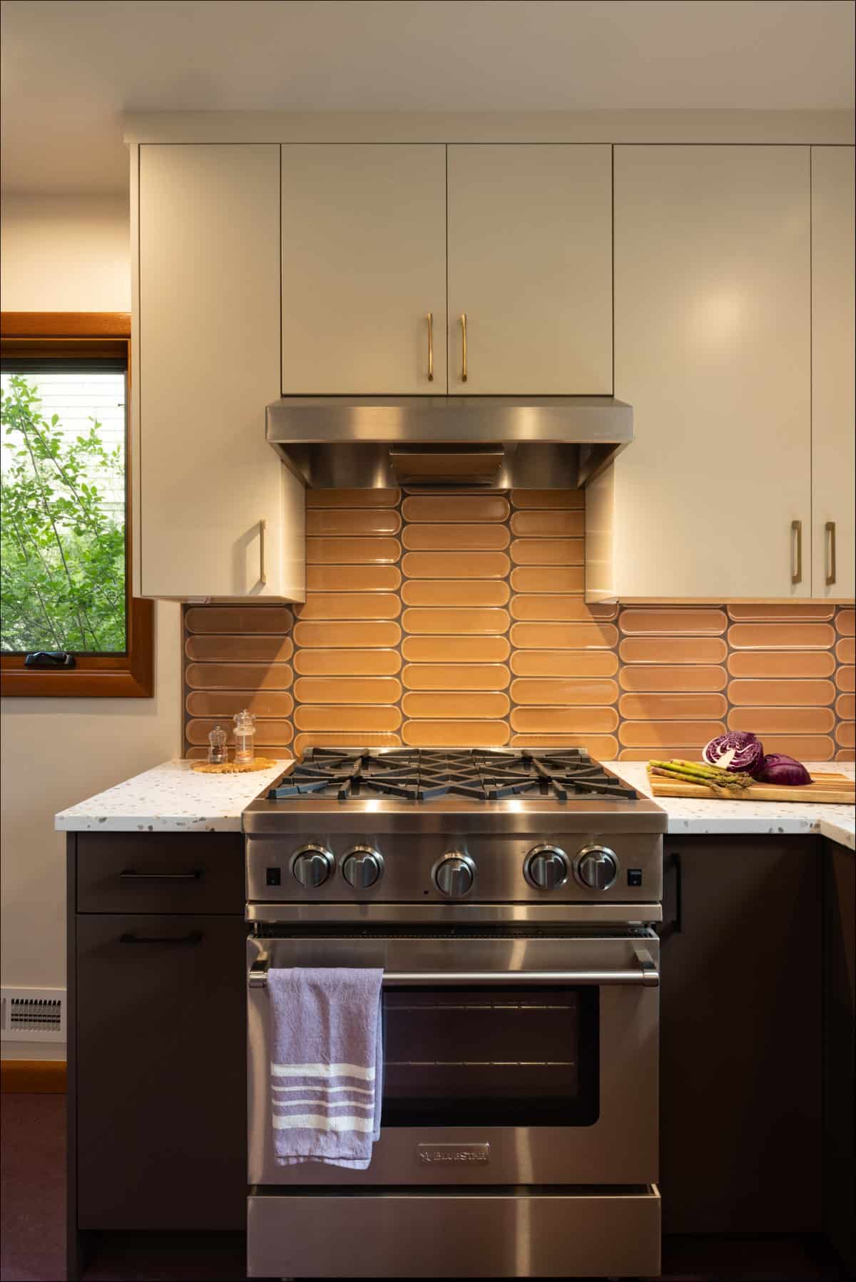 A modern kitchen with a stainless steel gas stove, beige tile backsplash, white upper cabinets, and a countertop with a towel hanging on the oven handle. A modern kitchen with a stainless steel gas stove, beige tile backsplash, white upper cabinets, and a countertop with a towel hanging on the oven handle.
