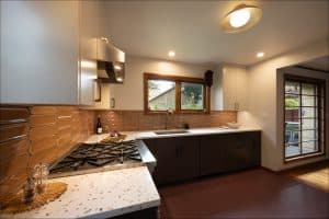 Modern kitchen with white upper cabinets, dark lower cabinets, a gas stove, a large sink beneath a window, brown tile backsplash, and overhead lighting.