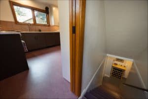 A view from a kitchen with brown cabinets and large windows into a stairway leading down to a room with a wine rack and framed pictures on the wall.